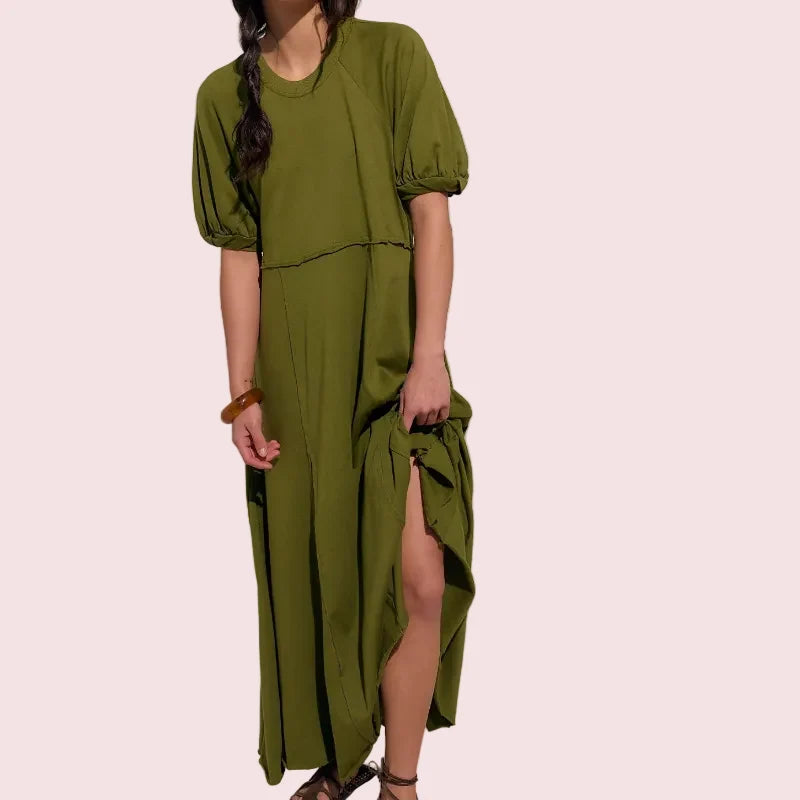 A woman modeling an olive green midi dress with puff sleeves, a high-low hem, and a side slit, styled with a mustard yellow headscarf and brown strappy sandals against a plain off-white studio background.