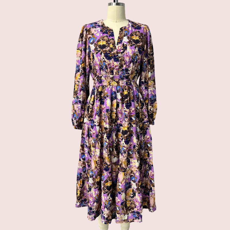 A long-sleeve midi dress with a purple, yellow, and black floral pattern, displayed on a mannequin in front of a plain white wall.