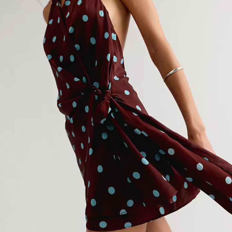 A woman wearing a sleeveless halter-neck mini dress in deep burgundy with light blue polka dots, tied at the waist. The photo is cropped from the chin down against a plain light gray studio background.