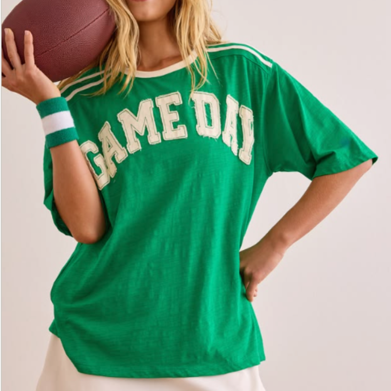 A woman wearing an oversized green 'GAME DAY' t-shirt with white varsity lettering, holding a brown football against her shoulder, set against a plain light beige background.