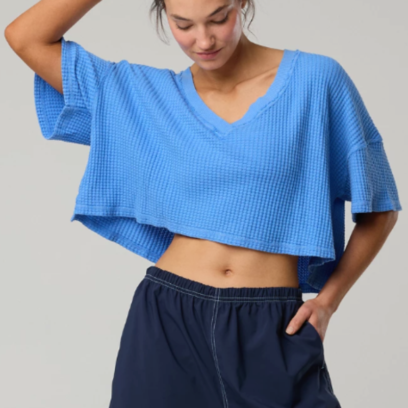A woman wearing a bright blue waffle-knit cropped t-shirt with short sleeves and a V-neck, paired with dark navy pants, against a solid light gray background.