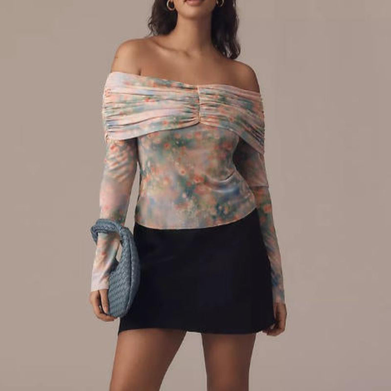 A woman models an off-the-shoulder long-sleeve top with a pastel multicolor floral print, paired with a black mini skirt and holding a blue textured clutch against a solid beige background.