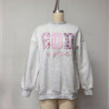 A light gray heather crewneck sweatshirt displayed on a mannequin torso. The front features a graphic spelling 'GOD' in pink gingham and floral patterned squares, with 'be good' written in pink script underneath.