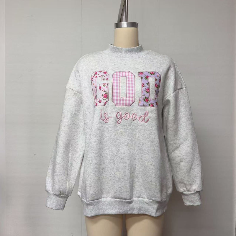 A light gray heather crewneck sweatshirt displayed on a mannequin torso. The front features a graphic spelling 'GOD' in pink gingham and floral patterned squares, with 'be good' written in pink script underneath.