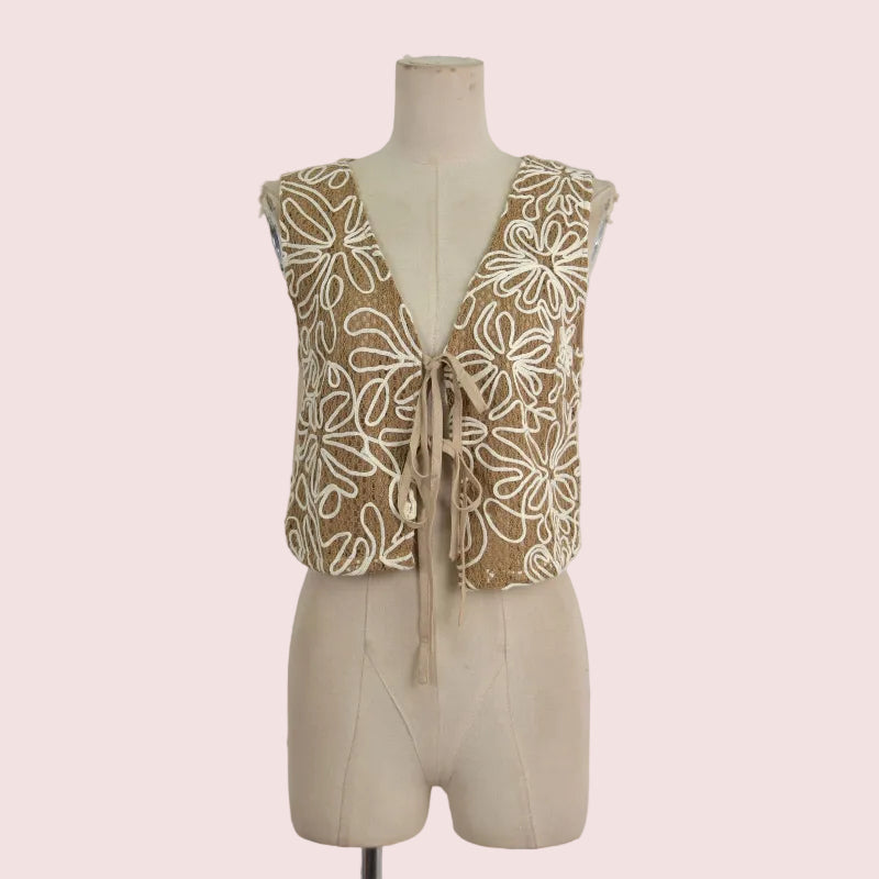 A sleeveless beige vest with a deep V-neck and tie-front closure, decorated with large cream floral embroidery, displayed on a beige dress form against a light gray background.