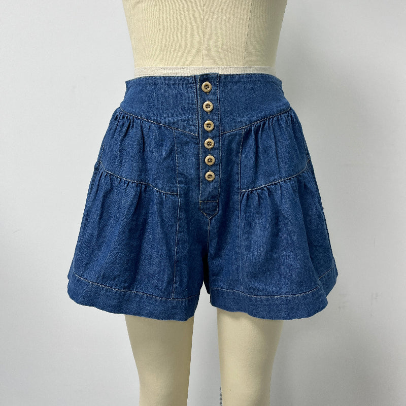 A pair of blue denim shorts with a high waist, tiered ruffle skirt, and a front button closure, displayed on a cream-colored dress form mannequin.