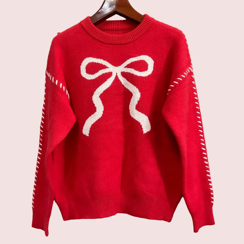 A bright red knit sweater hanging on a wooden hanger against a textured gray wall. The sweater features a large white bow motif on the chest and white dashed line details running down both sleeves.