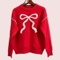 A bright red knit sweater hanging on a wooden hanger against a textured gray wall. The sweater features a large white bow motif on the chest and white dashed line details running down both sleeves.