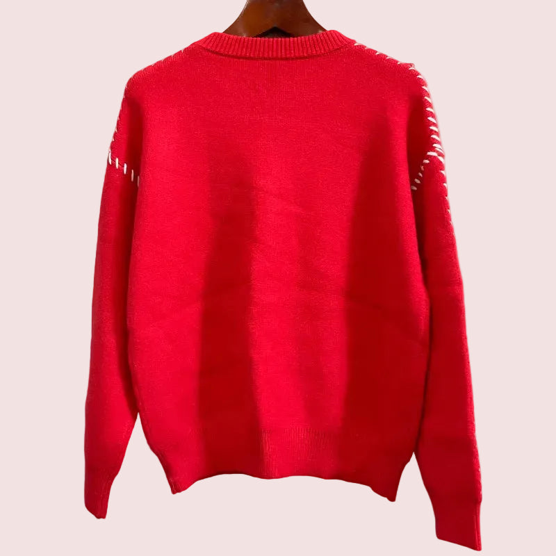 A bright red knit sweater hanging on a wooden hanger against a textured gray wall, featuring white contrast stitching along the shoulders and upper sleeves.