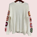 An off-white ribbed knit sweater with colorful crochet granny square patterns on the sleeves, hanging on a wooden hanger against a gray wall.