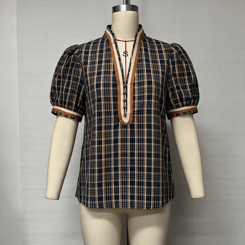A short-sleeve plaid blouse displayed on a cream mannequin torso against a plain light gray wall. The blouse has a dark navy and beige plaid pattern with orange-brown trim along the split neckline and sleeve cuffs.