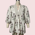 A cream-colored blouse with a purple and green floral print, featuring a black polka-dot V-neckline panel and puff sleeves, displayed on a mannequin against a solid pale pink background.