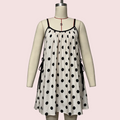 A sleeveless white slip dress with black polka dots and thin black straps, displayed on a cream mannequin against a pale pink background.