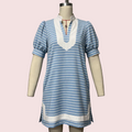 A short-sleeve tunic dress with light blue and white horizontal stripes, featuring a white V-neckline with a red drawstring detail, displayed on a cream-colored mannequin against a solid pale pink background.