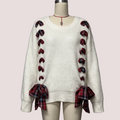 An off-white ribbed knit sweater displayed on a mannequin, featuring red and black plaid bow details down the front and tied at the cuffs.