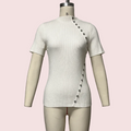 A short-sleeve ribbed knit top in off-white/cream displayed on a beige mannequin torso against a pale pink background. The top has an asymmetrical diagonal row of dark buttons running from the right shoulder area down the front, and a small size label 'S' is visible on the mannequin neck.