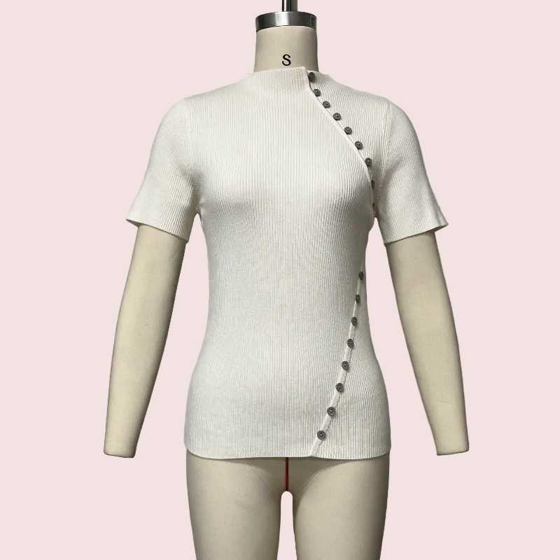 A short-sleeve ribbed knit top in off-white/cream displayed on a beige mannequin torso against a pale pink background. The top has an asymmetrical diagonal row of dark buttons running from the right shoulder area down the front, and a small size label 'S' is visible on the mannequin neck.