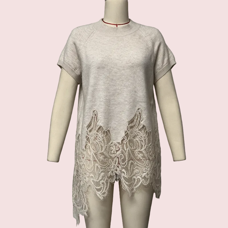 A light beige short-sleeve knit tunic top with a large beige lace panel at the hem, displayed on a mannequin against a pale pink background.