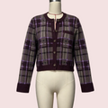 A cropped button-front cardigan sweater displayed on a cream mannequin torso against a pale pink background. The cardigan has a plaid pattern in dark purple, gray, and black with thin purple lines, ribbed dark purple collar, cuffs, and hem, and brown buttons.