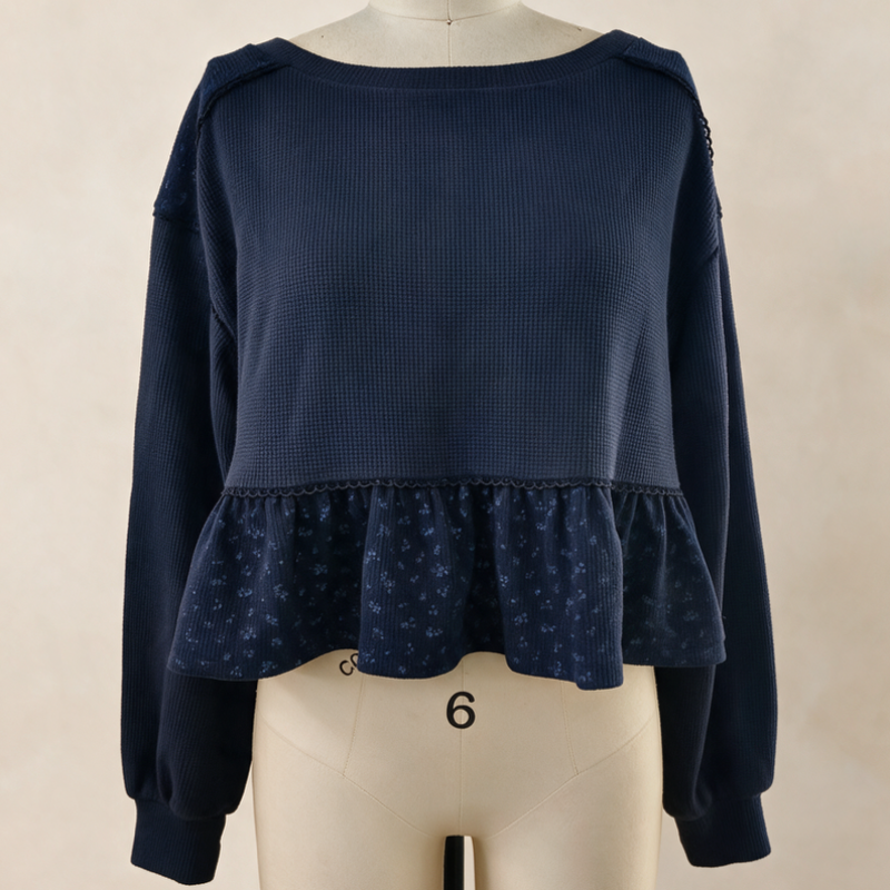 A navy blue cropped knit top with long sleeves, a wide boat neckline, and a ruffled peplum hem, displayed on a cream mannequin against a neutral beige background.