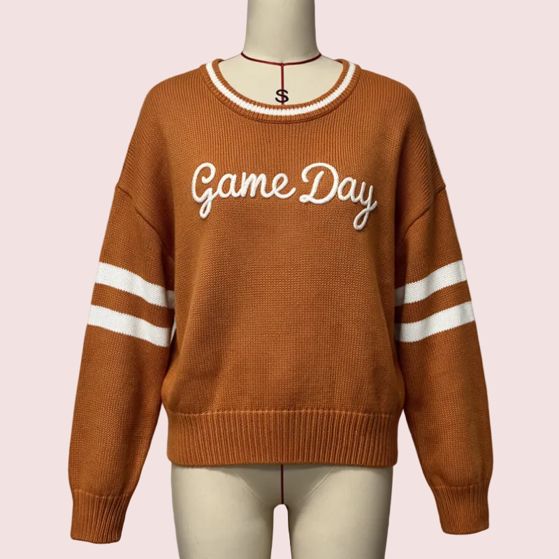 A rust-orange knit sweater displayed on a mannequin, featuring 'Game Day' embroidered in white script across the chest and white stripes on the sleeves.